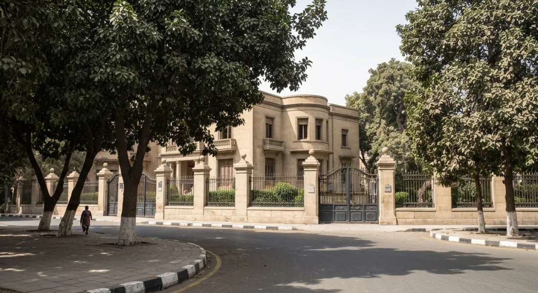 una calle tranquila y curva de Garden City al mediodía, ficus sombreando una villa Art Deco tras puertas de estilo embajada y barreras de seguridad